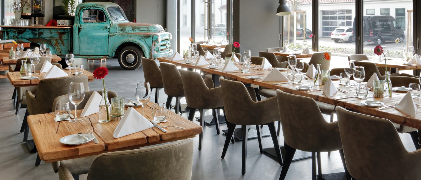 Ein modernes Restaurant mit Holztischen, eleganten Stühlen und einem alten blauen Truck als Dekoration. Große Fenster lassen viel Licht herein., © V8 Hotel, Frank Hoppe Ein modernes Restaurant mit Holztischen, eleganten Stühlen und einem alten blauen Truck als Dekoration. Große Fenster lassen viel Licht herein., © V8 Hotel, Frank Hoppe