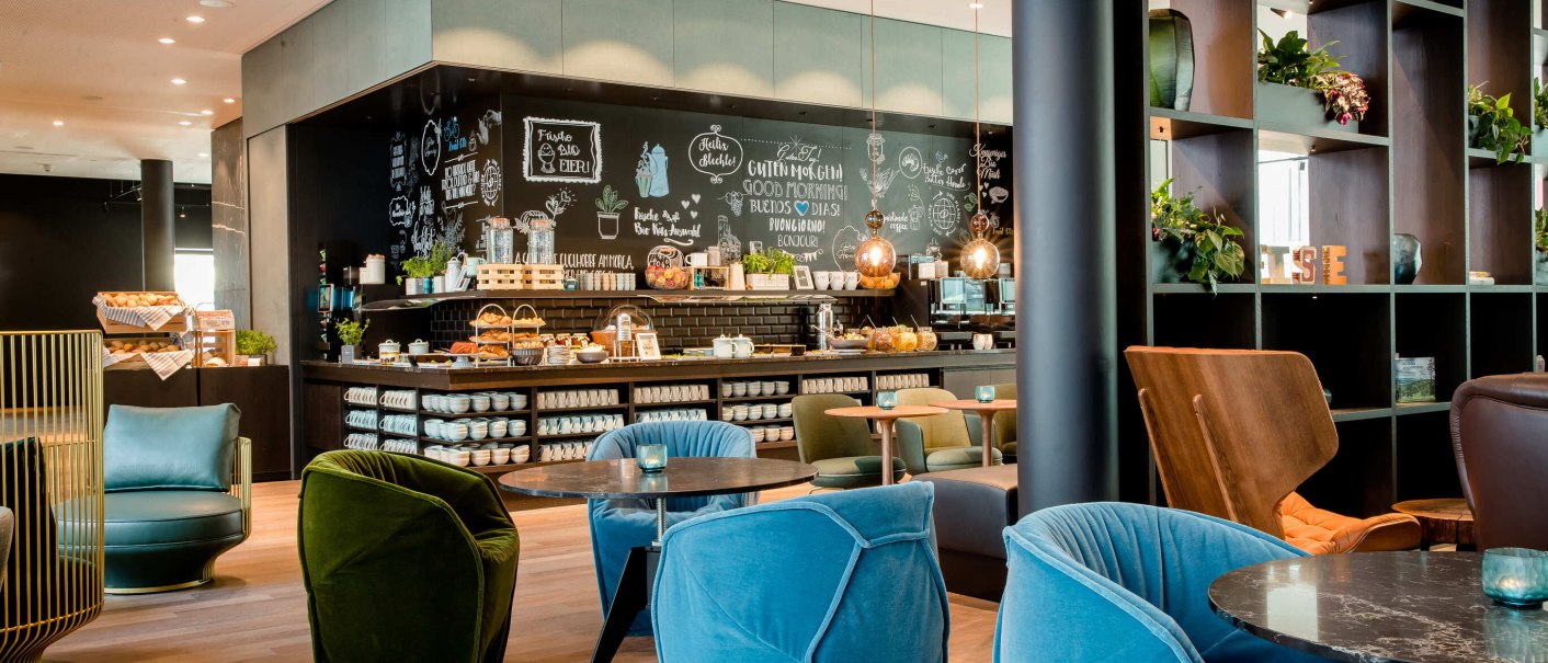 Moderne Lobby mit bunten Sesseln, Tischen und einer Kaffeebar. Die Wand ist mit Kreidezeichnungen dekoriert. Pflanzen und Regale ergänzen das Ambiente., © Motel One - Stuttgart Hauptbahnhof