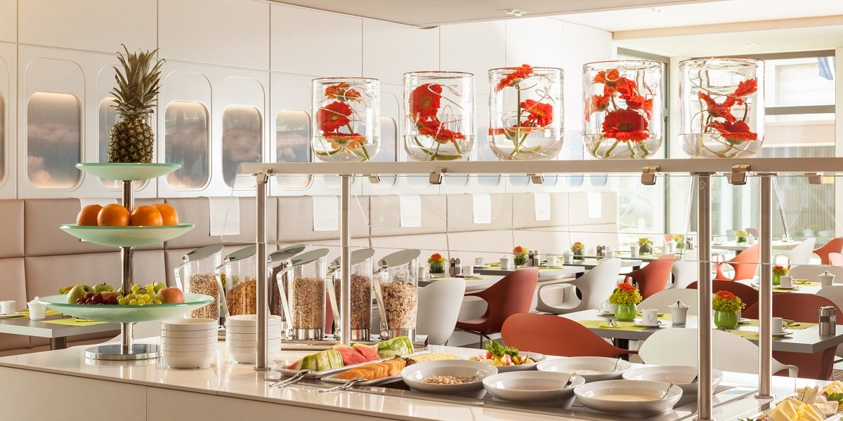 Modernes Restaurant mit stilvollem Buffet. Obst, Müsli und Blumenarrangements in Gläsern. Helle, einladende Atmosphäre mit eleganten Sitzgelegenheiten., © Essential by Dorint