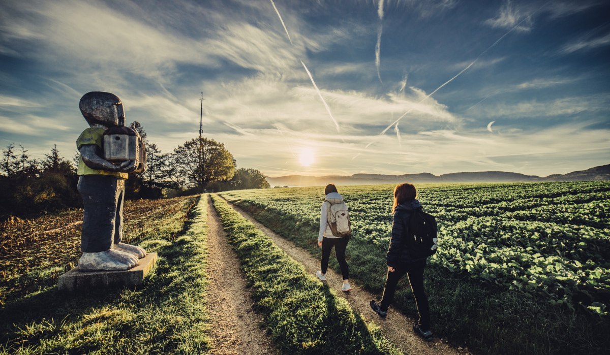 Zwei Personen wandern bei Sonnenaufgang auf einem Feldweg. Links steht eine große Skulptur. Der Himmel ist klar mit einigen Wolkenstreifen., © Landratsamt Ostalbkreis