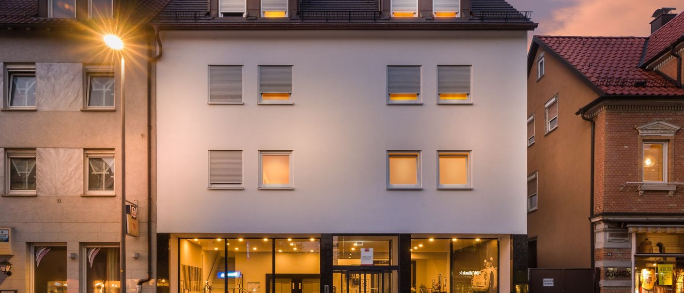 Modernes Gebäude mit großen Fenstern und beleuchtetem Innenraum, umgeben von älteren Häusern. Ein Auto parkt davor, Straßenlaterne leuchtet., © Novum Hospitality