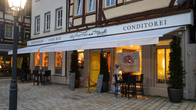 Außenansicht eines Restaurants mit Café und Konditorei. Beleuchtete Fenster und Tische im Freien bei Dämmerung. Schriftzug 'AlbThermen am Markt' sichtbar.