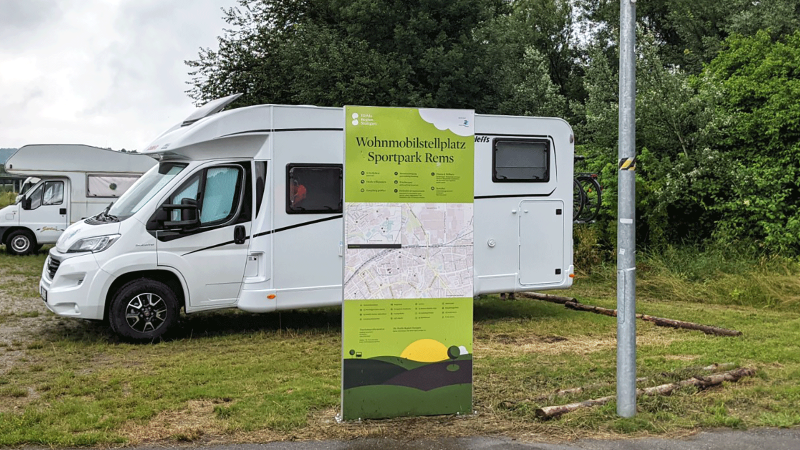 Zwei Wohnmobile parken auf einem Stellplatz im Sportpark Rems. Ein großes Schild informiert über den Standort. Im Hintergrund sind Bäume zu sehen., © Stadtverwaltung Schorndorf