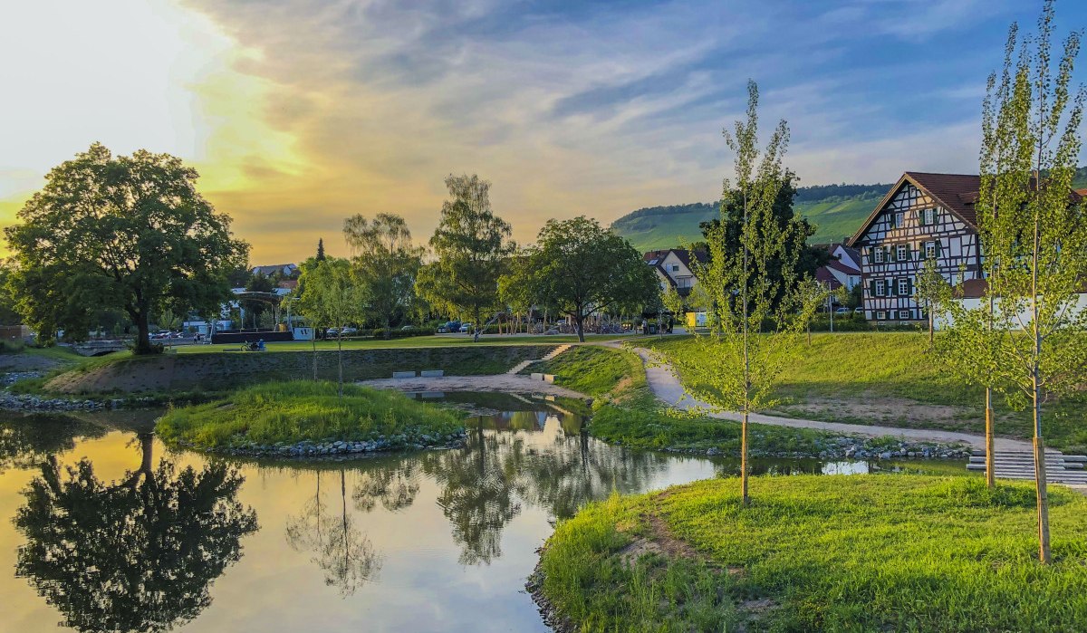 Idyllische Landschaft mit Fluss, Bäumen und Fachwerkhäusern bei Sonnenuntergang in Weinstadt. Der Himmel ist blau mit gelben und orangen Tönen., © Stadt Weinstadt Idyllische Landschaft mit Fluss, Bäumen und Fachwerkhäusern bei Sonnenuntergang in Weinstadt. Der Himmel ist blau mit gelben und orangen Tönen., © Stadt Weinstadt