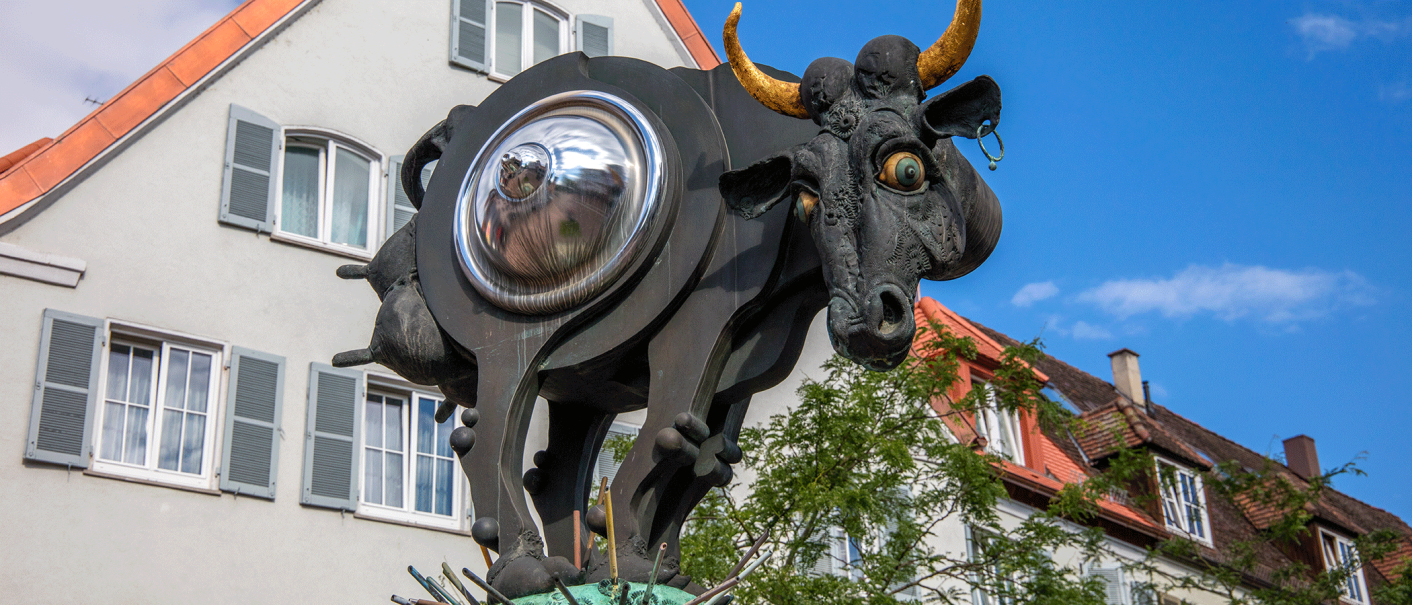 Surreale Kuhskulptur mit metallischem Bauch und goldenen Hörnern vor einem Gebäude mit roten Dachziegeln., © Stuttgart-Marketing GmbH