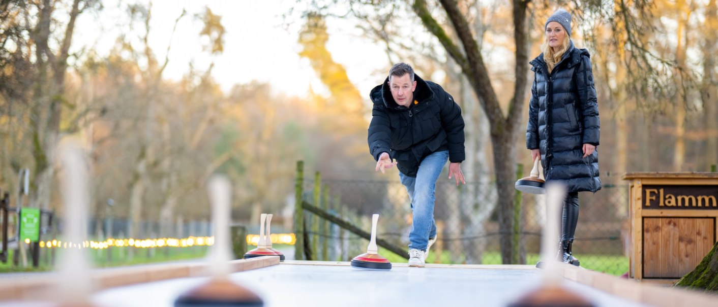 Ein Mann und eine Frau spielen Eisstockschießen im Freien. Der Mann wirft gerade einen Eisstock, während die Frau zuschaut. Bäume im Hintergrund., © Katzenbacher Hof Ein Mann und eine Frau spielen Eisstockschießen im Freien. Der Mann wirft gerade einen Eisstock, während die Frau zuschaut. Bäume im Hintergrund., © Katzenbacher Hof