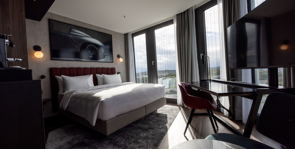 Modernes Eckzimmer mit großem Bett, Schreibtisch, Panoramafenstern und stilvollem Dekor. Ein Bild eines Autos hängt an der Wand., © Radisson Hotel Group