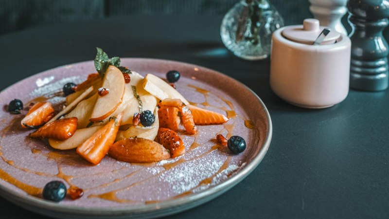 Ein Teller mit geschnittenen Früchten, Beeren und Sirup. Daneben eine Zuckerdose und ein Salzstreuer auf einem dunklen Tisch., © Lupin, Stuttgart Ein Teller mit geschnittenen Früchten, Beeren und Sirup. Daneben eine Zuckerdose und ein Salzstreuer auf einem dunklen Tisch., © Lupin, Stuttgart