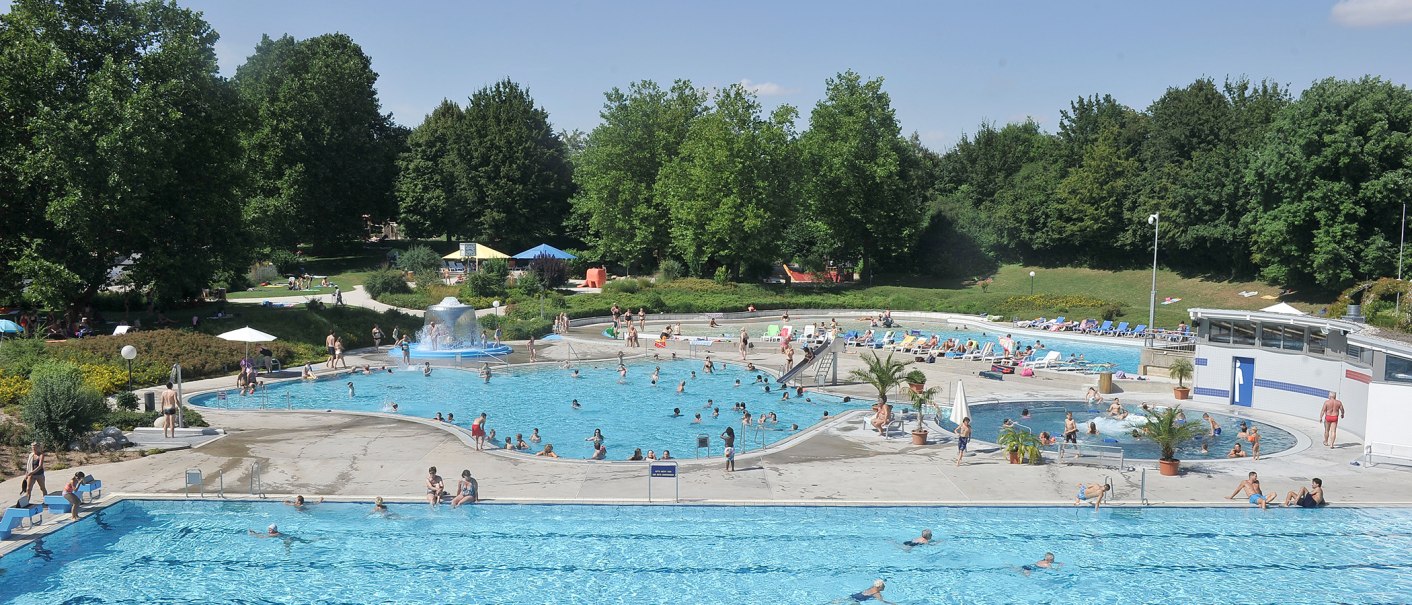 Ein belebtes Freibad mit mehreren Pools, vielen Besuchern und umgeben von grünen Bäumen. Menschen schwimmen und entspannen auf Liegestühlen., © Werner Kuhnle Ein belebtes Freibad mit mehreren Pools, vielen Besuchern und umgeben von grünen Bäumen. Menschen schwimmen und entspannen auf Liegestühlen., © Werner Kuhnle