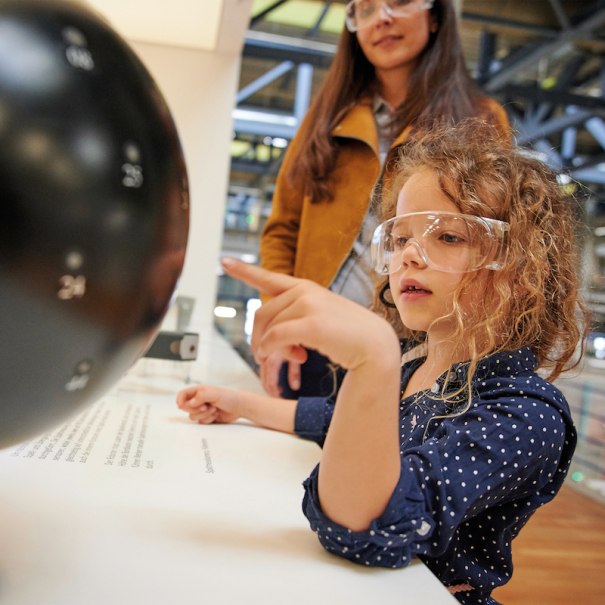 Ein Kind mit Schutzbrille erkundet interessiert ein Exponat, w&auml;hrend eine Frau im Hintergrund steht. Die Szene spielt in einem modernen Ausstellungsraum., &copy; Mercedes-Benz AG