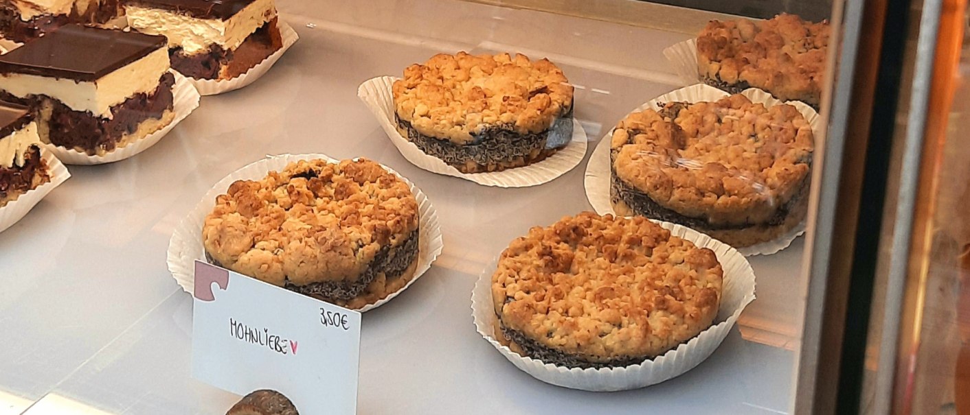 In einer Vitrine liegen Mohnkuchen und Schokoladenkuchen. Ein Schild zeigt den Preis von 3,50€ für den Mohnkuchen an., © SMG