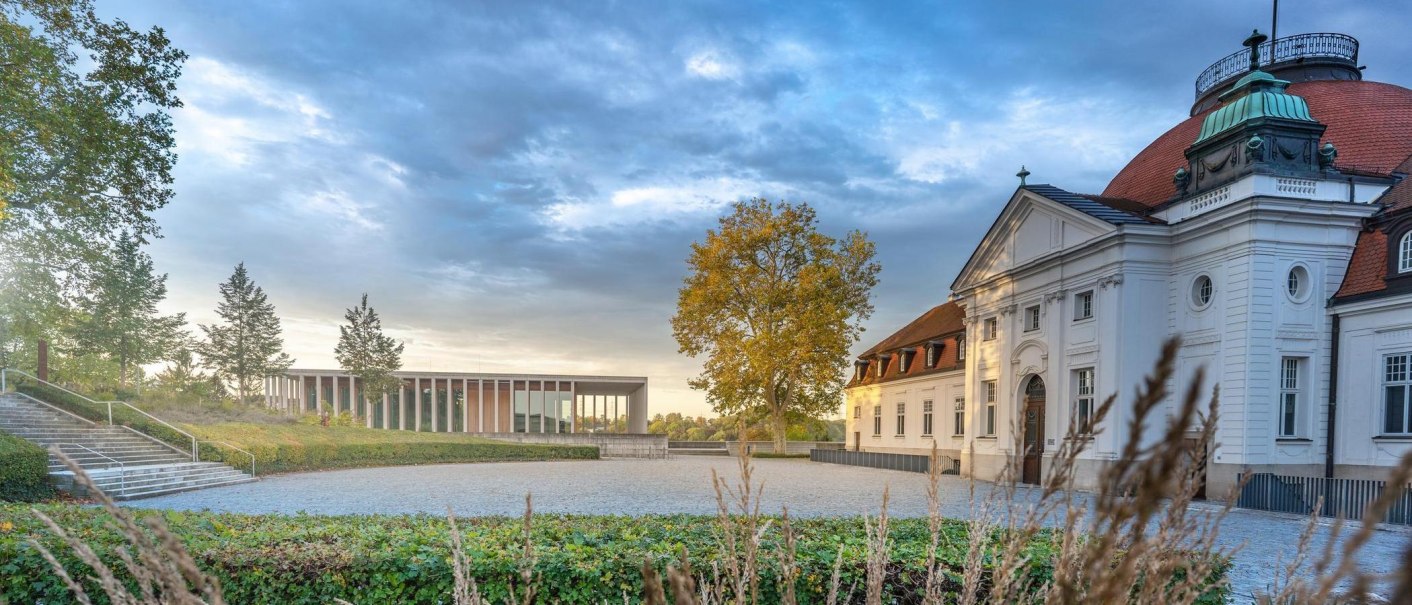 Ein historisches Gebäude mit rotem Dach und einem modernen Anbau im Hintergrund, umgeben von herbstlichen Bäumen und einem gepflasterten Platz., © SMG, Martina Denker Ein historisches Gebäude mit rotem Dach und einem modernen Anbau im Hintergrund, umgeben von herbstlichen Bäumen und einem gepflasterten Platz., © SMG, Martina Denker