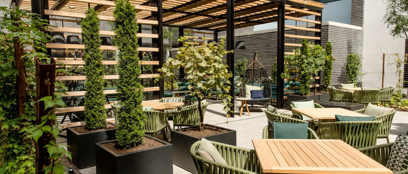 Moderne Terrasse mit Holztischen, grünen Stühlen und Pflanzen. Eine Pergola spendet Schatten, während Kletterpflanzen und Bäume für eine grüne Atmosphäre sorgen., © Motel One - Stuttgart Hauptbahnhof