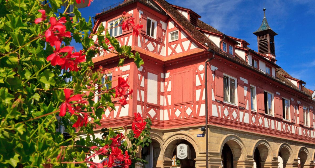 Das Alte Rathaus in Waiblingen, ein Fachwerkgebäude mit roten und weißen Details, umgeben von blühenden roten Blumen unter blauem Himmel., © WTM GmbH, Kai Koepf