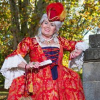 Eine Person in einem aufwendigen, roten historischen Kost&uuml;m posiert l&auml;chelnd neben einer Steins&auml;ule im Freien. Herbstliche B&auml;ume im Hintergrund., &copy; &copy; T&E, Reiner Pfisterer
