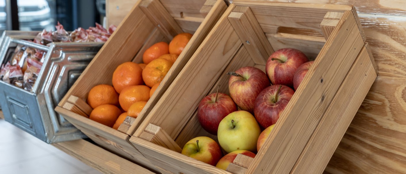 Holzkisten mit frischen &Auml;pfeln und Orangen, daneben ein Metallbeh&auml;lter mit verpackten Snacks in einem Fr&uuml;hst&uuml;cksbereich., &copy; a2 HOTELS