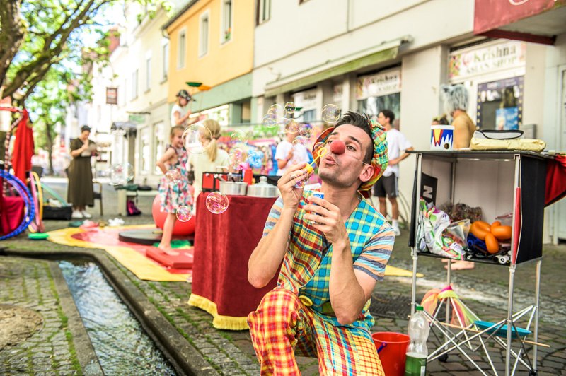 Ein Clown kniet in der Fußgängerzone und macht Seifenblasen. Im Hintergrund sind Kinder zu sehen., © Marcus Gauß Ein Clown kniet in der Fußgängerzone und macht Seifenblasen. Im Hintergrund sind Kinder zu sehen., © Marcus Gauß