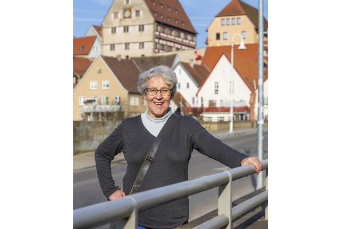 Eine ältere Frau steht lächelnd auf einer Brücke in Besigheim. Im Hintergrund sind malerische Fachwerkhäuser zu sehen., © Stadt Besigheim Eine ältere Frau steht lächelnd auf einer Brücke in Besigheim. Im Hintergrund sind malerische Fachwerkhäuser zu sehen., © Stadt Besigheim