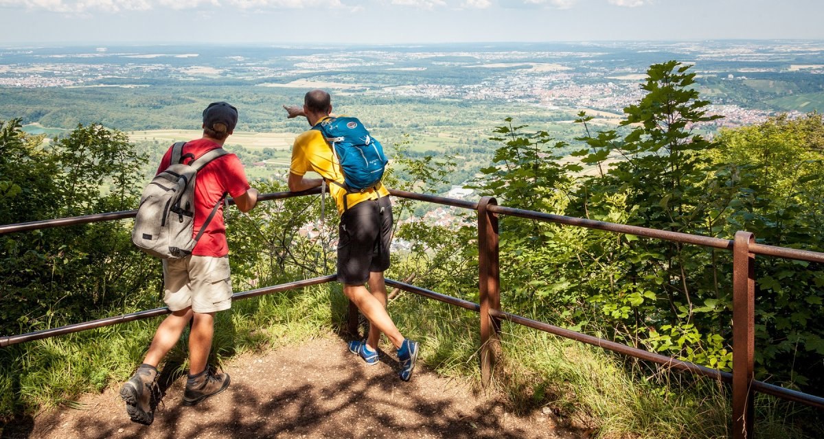 Zwei Wanderer mit Rucksäcken stehen an einem Geländer und blicken auf eine weite, grüne Landschaft unter einem blauen Himmel mit Wolken., © hochgehberge Zwei Wanderer mit Rucksäcken stehen an einem Geländer und blicken auf eine weite, grüne Landschaft unter einem blauen Himmel mit Wolken., © hochgehberge