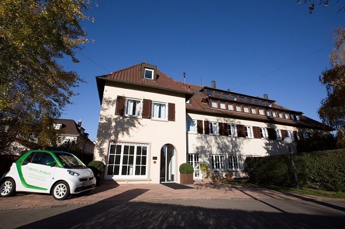 Ein zweist&ouml;ckiges Hotel mit braunen Fensterl&auml;den, umgeben von B&auml;umen. Ein kleines Auto steht davor. Der Himmel ist klar und blau., &copy; Hotel B&uuml;rkle