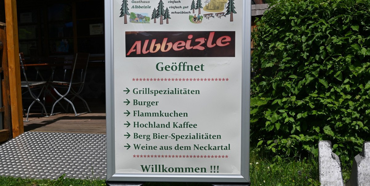Schild des Gasthauses 'Albbeizle' mit Angeboten wie Grillspezialitäten, Burger, Flammkuchen, Kaffee, Bier und Wein. Im Hintergrund sind Tische zu sehen.