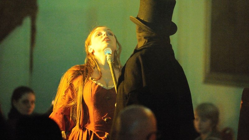 Eine Frau in einem roten Kleid steht vor einem Mann in schwarzem Mantel und Zylinder. Die Szene wirkt dramatisch und ist in warmem Licht gehalten., © Gruseldinner GmbH & Co. KG Eine Frau in einem roten Kleid steht vor einem Mann in schwarzem Mantel und Zylinder. Die Szene wirkt dramatisch und ist in warmem Licht gehalten., © Gruseldinner GmbH & Co. KG