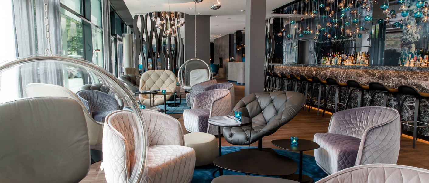 Moderne Lobby mit eleganten Sesseln, runden Tischen und einer stilvollen Bar. Hängelampen und große Fenster schaffen eine einladende Atmosphäre., © Motel One Stuttgart-Bad Cannstatt