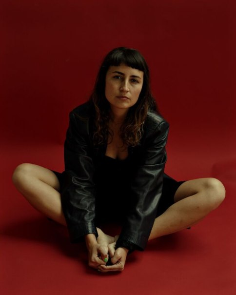 Eine Person in schwarzer Lederjacke sitzt mit verschränkten Beinen auf einem roten Hintergrund und schaut in die Kamera., © Stadt Böblingen - Amt für Kultur