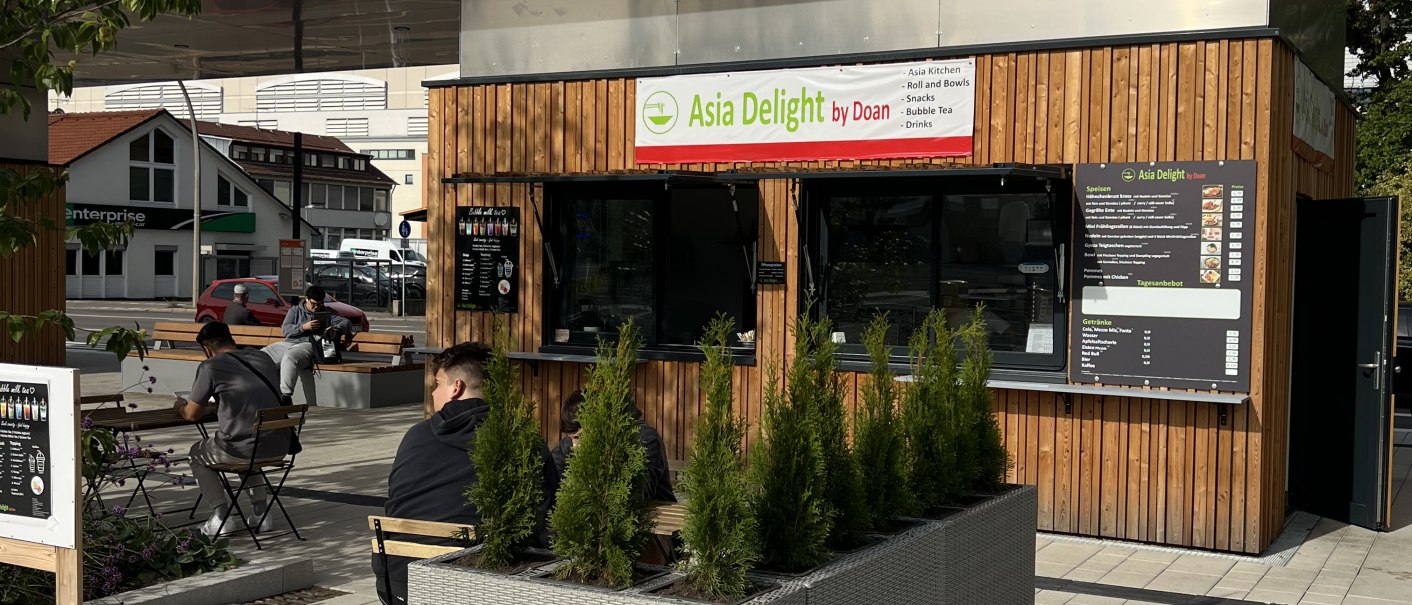 Kleiner Kiosk 'Asia Delight by Doan' mit Holzverkleidung und Sitzbereich im Freien. Menschen sitzen an Tischen, umgeben von Pflanzen., © Asia Delight GmbH