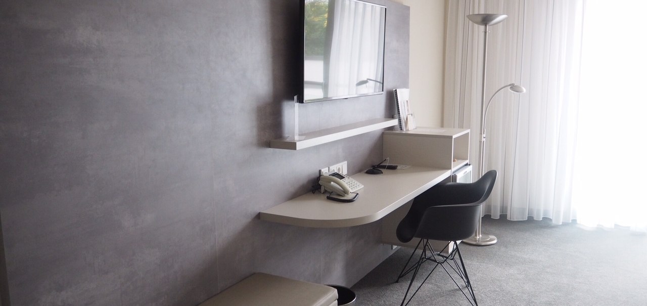 Moderne Hotelsuite mit grauem Schreibtisch, schwarzem Stuhl, Wandfernseher und Stehlampe vor einem großen Fenster mit weißen Vorhängen., © Hotel Fuchsen Moderne Hotelsuite mit grauem Schreibtisch, schwarzem Stuhl, Wandfernseher und Stehlampe vor einem großen Fenster mit weißen Vorhängen., © Hotel Fuchsen