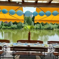 Gedeckte Tische unter gelben Markisen, dekoriert mit blauen Luftballons, mit Blick auf einen See und grüne Bäume im Hintergrund., © Gaststätte Goldbachsee