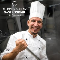 Ein Koch in wei&szlig;er Uniform und M&uuml;tze l&auml;chelt in einer modernen K&uuml;che. Er h&auml;lt einen gro&szlig;en L&ouml;ffel. Oben links ist das Logo von Mercedes-Benz Gastronomie zu sehen., &copy; Mercedes-Benz AG