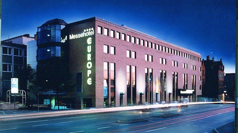 Beleuchtetes Messehotel Europe bei Nacht, mit leuchtendem Schriftzug und vorbeifahrenden Autos auf der Straße., © Messehotel Europe Beleuchtetes Messehotel Europe bei Nacht, mit leuchtendem Schriftzug und vorbeifahrenden Autos auf der Straße., © Messehotel Europe