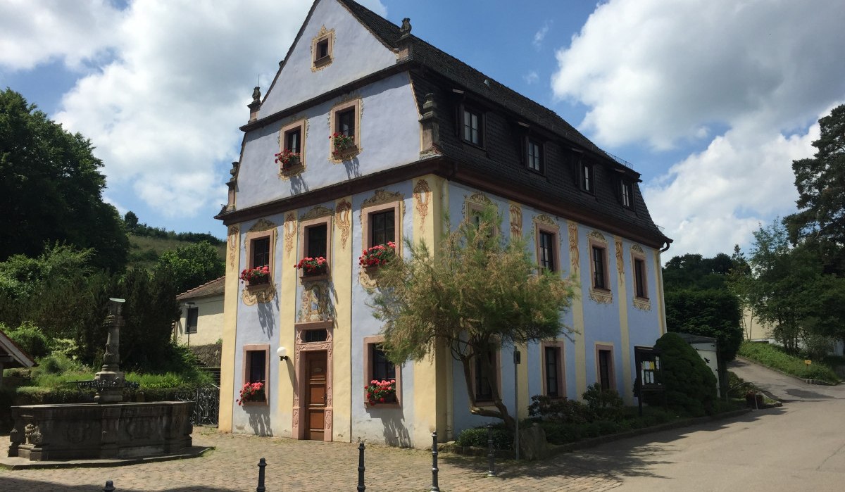 Historisches Gebäude mit verzierten Fenstern und Blumenkästen, daneben ein Brunnen, umgeben von Bäumen und einem gepflasterten Weg., © www.pro-cycl.de