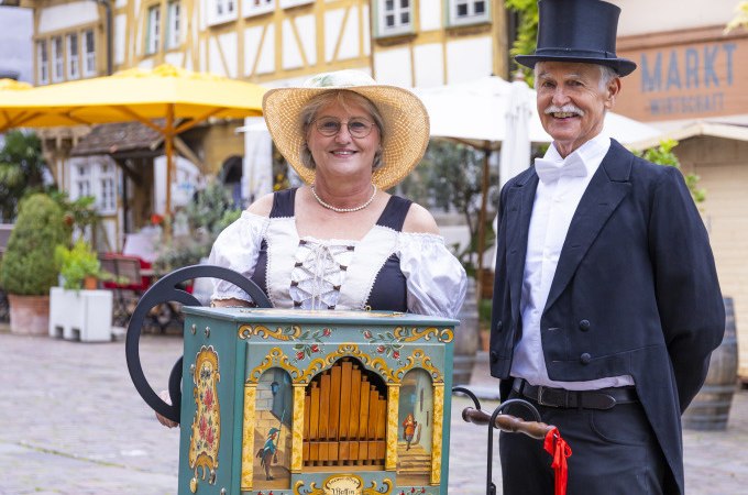 Ein Paar in historischer Kleidung steht mit einer Drehorgel auf einem malerischen Platz. Im Hintergrund sind Fachwerkhäuser und ein Marktstand zu sehen., © Stadt Besigheim Ein Paar in historischer Kleidung steht mit einer Drehorgel auf einem malerischen Platz. Im Hintergrund sind Fachwerkhäuser und ein Marktstand zu sehen., © Stadt Besigheim