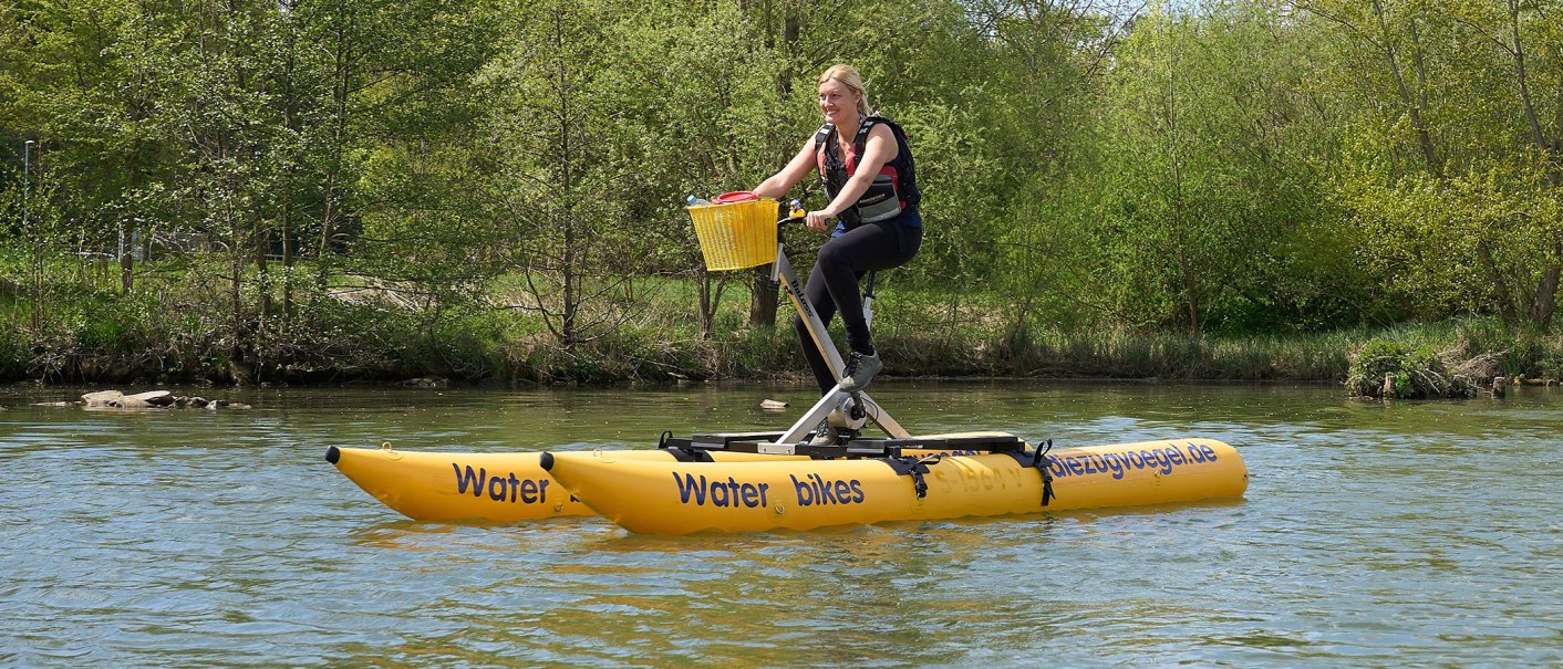 Eine Person fährt auf einem gelben Waterbike über einen Fluss, umgeben von grünen Bäumen. Das Waterbike hat zwei Schwimmkörper und einen Korb vorne., © Foto LKZ - Fotograf Andreas Becker