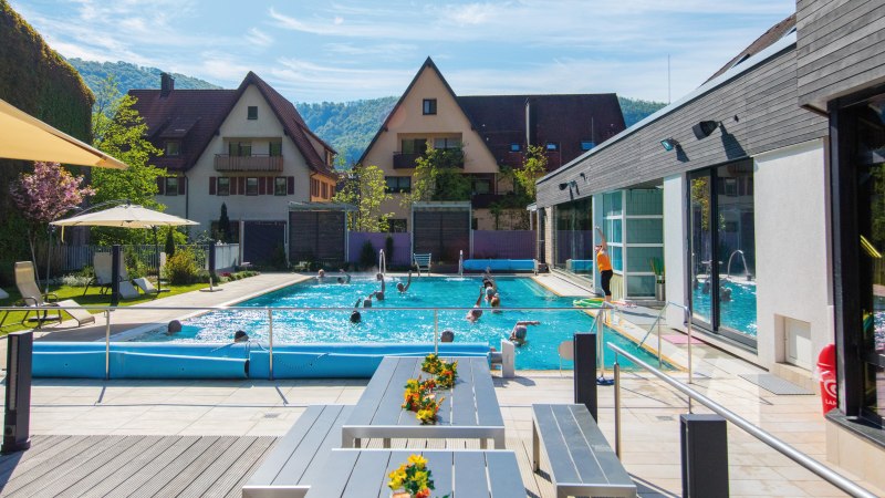 Thermalbad mit Pool, Liegestühlen und Sonnenschirmen. Menschen schwimmen im Wasser, im Hintergrund sind Häuser und grüne Hügel zu sehen., © SMG Achim Mende
