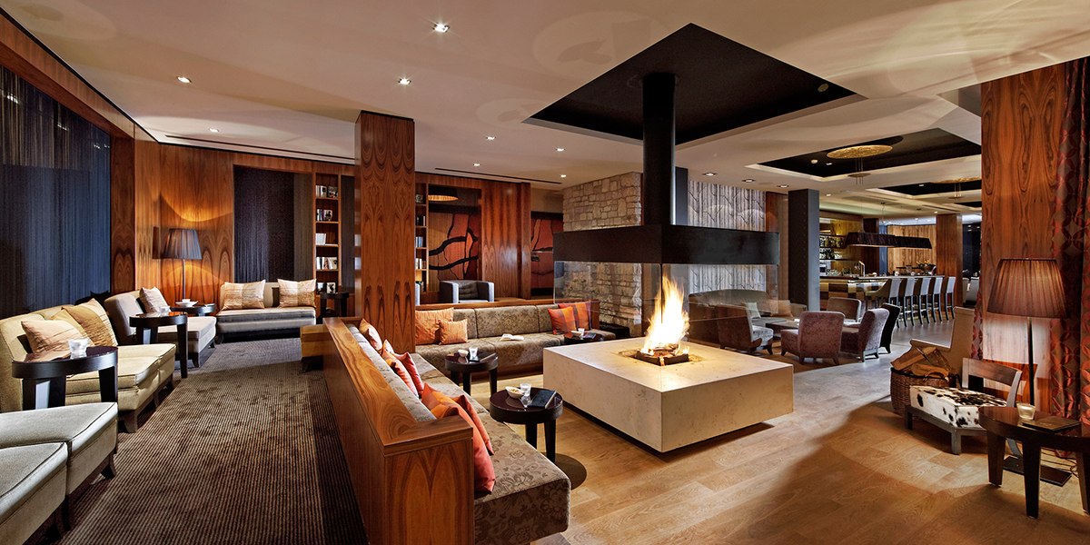 Elegante Lounge mit zentralem Kamin, gemütlichen Sofas und einer Bar im Hintergrund. Holzwände und warme Beleuchtung schaffen eine einladende Atmosphäre., © Parkhotel Stuttgart Messe-Airport