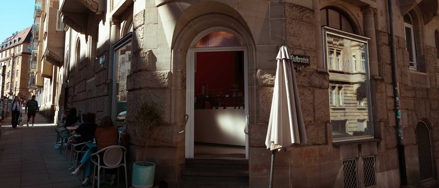 Ein Café in der Gutbrodstraße mit Sitzgelegenheiten im Freien. Menschen sitzen an Tischen, während andere die Straße entlanggehen., © SMG Ein Café in der Gutbrodstraße mit Sitzgelegenheiten im Freien. Menschen sitzen an Tischen, während andere die Straße entlanggehen., © SMG