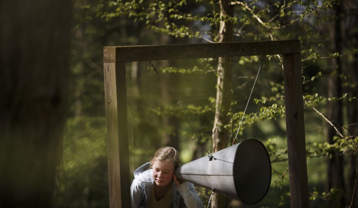Ein Mädchen lauscht einem großen Metalltrichter, der an einem Holzrahmen im Wald befestigt ist. Die Umgebung ist grün und bewaldet., © Natur.Nah. Schönbuch & Heckengäu Ein Mädchen lauscht einem großen Metalltrichter, der an einem Holzrahmen im Wald befestigt ist. Die Umgebung ist grün und bewaldet., © Natur.Nah. Schönbuch & Heckengäu