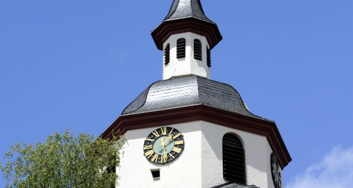 Ein Kirchturm mit Uhr und Wetterhahn ragt in den blauen Himmel. Ein Baum und ein Ziegeldach sind im Vordergrund sichtbar., &copy; Natur.Nah. Sch&ouml;nbuch & Heckeng&auml;u