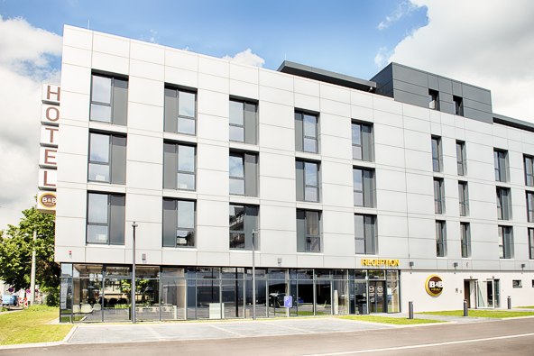 Modernes Hotelgebäude mit Glasfassade, Beschilderung 'Hotel' und 'Rezeption', umgeben von Grünflächen., © B&B Hotel Stuttgart-Neckarhafen