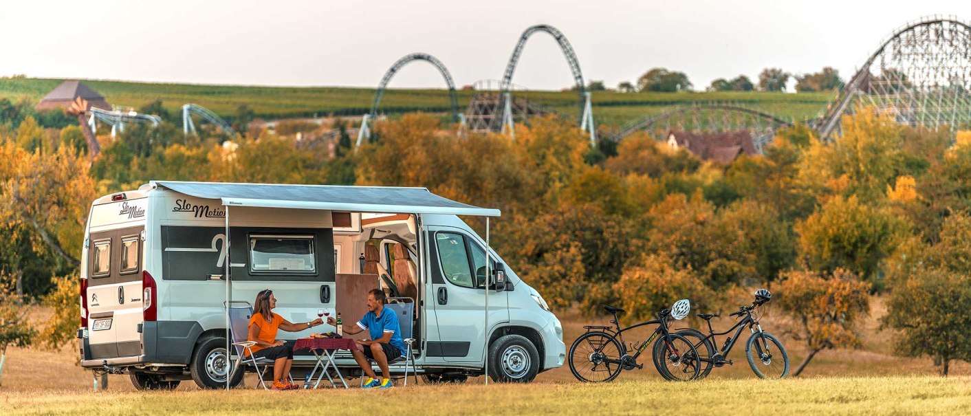 Wohnmobil mit Markise auf Wiese, zwei Personen am Tisch, Fahrräder daneben, Achterbahnen im Hintergrund, herbstliche Landschaft., © Erlebnispark Tripsdrill GmbH & Co. KG