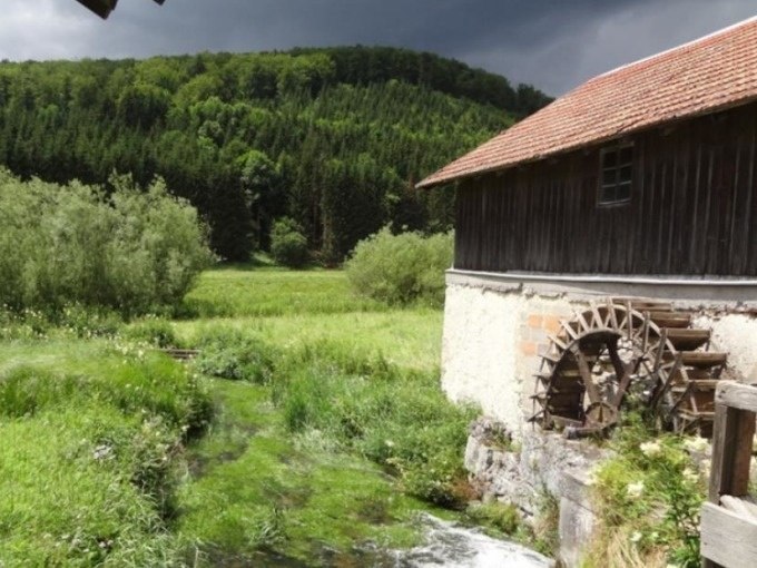 Eine historische Mühle mit Wasserrad an einem klaren Bach, umgeben von grüner Landschaft und einem bewaldeten Hügel im Hintergrund., © SwabianTravel Eine historische Mühle mit Wasserrad an einem klaren Bach, umgeben von grüner Landschaft und einem bewaldeten Hügel im Hintergrund., © SwabianTravel