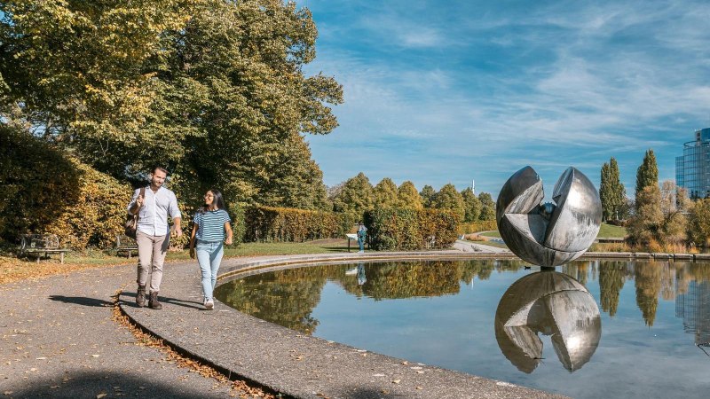 Ein Paar spaziert im Wartbergpark entlang eines Teiches mit einer gro&szlig;en Skulptur und einem modernen Geb&auml;ude im Hintergrund., &copy; Stuttgart-Marketing GmbH, Sarah Schmid