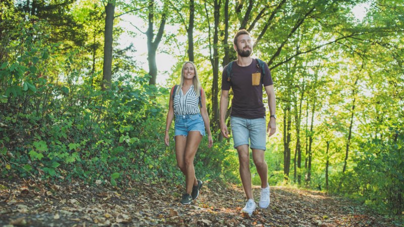 Ein Paar genie&szlig;t eine Wanderung auf einem Waldweg im Remstal, umgeben von &uuml;ppigem Gr&uuml;n und Sonnenlicht.