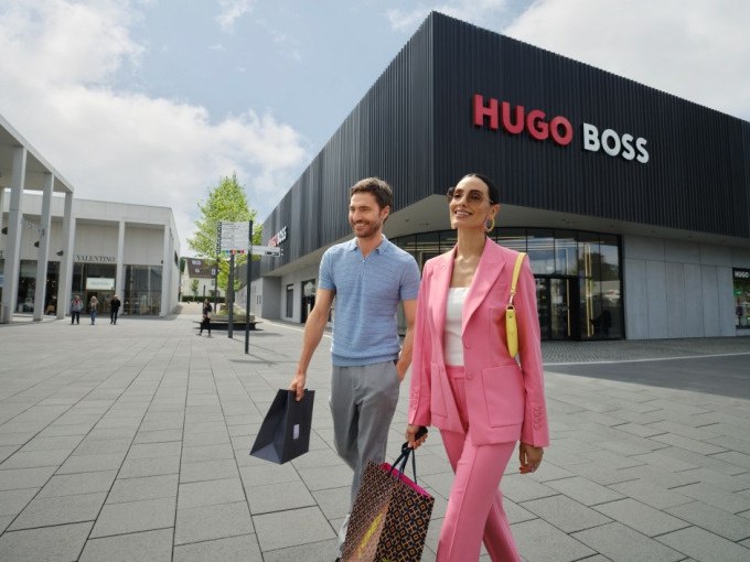 Zwei Personen mit Einkaufstaschen gehen l&auml;chelnd vor einem Hugo Boss Gesch&auml;ft in einem modernen Einkaufszentrum spazieren., &copy; Schlienz-Tours GmbH &amp; Co.KG