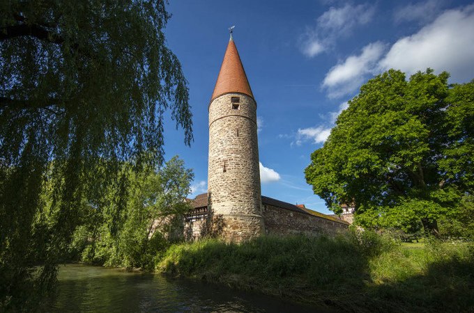 Ein mittelalterlicher Turm mit rotem Dach steht malerisch neben einem Fluss, umgeben von &uuml;ppigem Gr&uuml;n und einer alten Steinmauer., &copy; Stadtverwaltung Weil der Stadt
