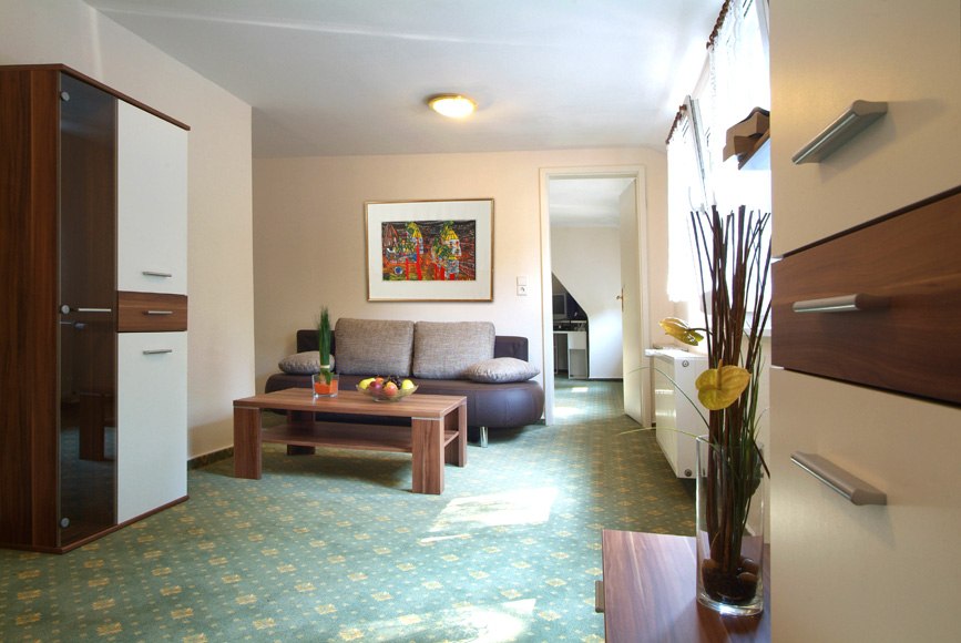 Helles Wohnzimmer mit Sofa, Couchtisch und Schrank. Ein Gemälde hängt an der Wand, und durch die Tür ist eine Küche sichtbar., © Kulinarium an der Glems