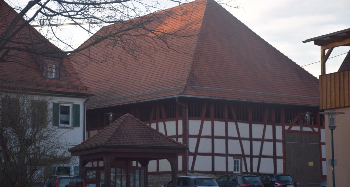 Ein großes Fachwerkgebäude mit rotem Ziegeldach, umgeben von geparkten Autos und einem Baum im Vordergrund., © Land der 1000 Hügel - Kraichgau-Stromberg Ein großes Fachwerkgebäude mit rotem Ziegeldach, umgeben von geparkten Autos und einem Baum im Vordergrund., © Land der 1000 Hügel - Kraichgau-Stromberg